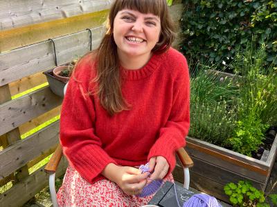 Maria sidder ved et havebord, mens hun strikker fra et lilla garnnøgle. På bordet står et keramikkrus med kaffe.